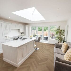 A modern white kitchen and living area featuring Bridle Oak herringbone flooring under bright natural light.